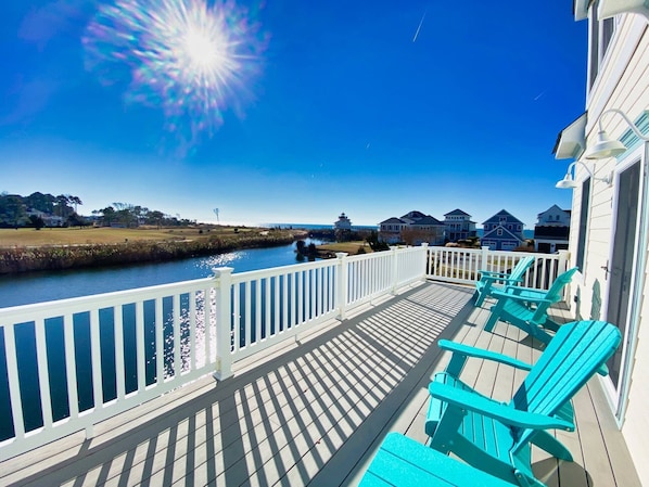 A wooden deck with a white railing, turquoise Adirondack chairs, and a view of a river and houses.