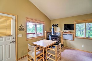 A cozy room with a wooden table and chairs, a stone fireplace, a television, and large windows with a view of trees. - Trego Cabin w/ Mtn Views, Easy Access to Lake (Trego)