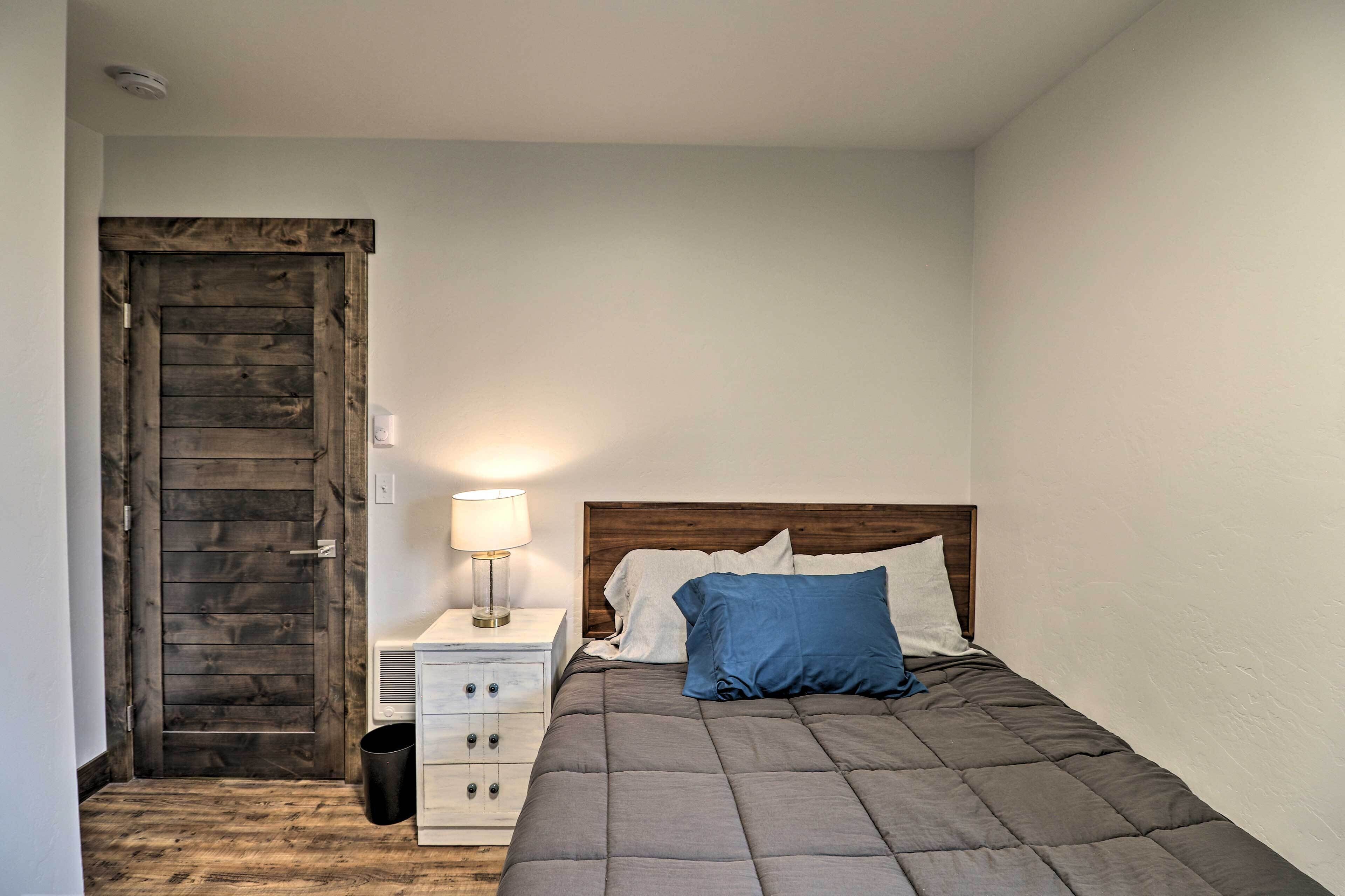 A bedroom with a wooden door, a bedside table with a lamp, a bed with a quilted blanket and pillows, and a white nightstand.