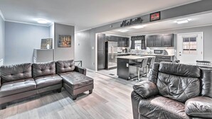 Un salon moderne avec un canapé d’angle et un fauteuil en cuir, une cuisine avec des meubles sombres et des appareils en acier inoxydable, et un coin repas avec un bar.