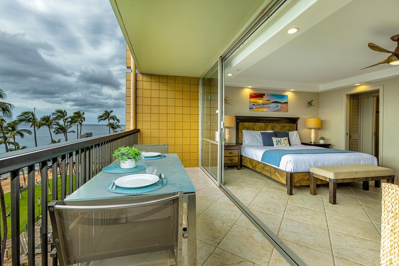 A balcony with a table set for two, a bed with a view of the ocean, and a ceiling fan.