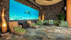 A modern lounge with a large wall-mounted TV, stone walls, and a variety of seating options including sofas and chairs. - Mana Kai #614 2 Bedroom Condo (Kihei)