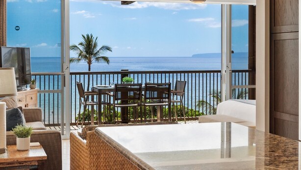 A modern living room with a large window overlooking the sea, a dining area with outdoor furniture, and a kitchen with a marble countertop. - Mana Kai #614 2 Bedroom Condo (Kihei)
