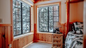A cozy room with wooden paneled walls, large windows with a view of snow-covered trees, a wooden dresser, and a bed with a forest-themed bedspread.