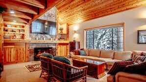 A cozy living room with a fireplace, wooden ceiling, and a large window with a snowy view.
