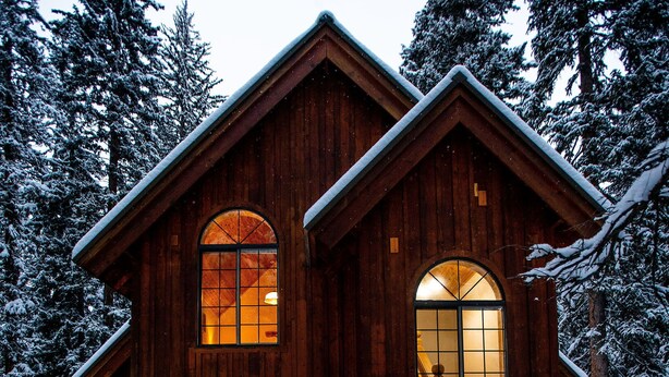 A wooden cabin with a staircase leading to the entrance, surrounded by snow-covered trees.