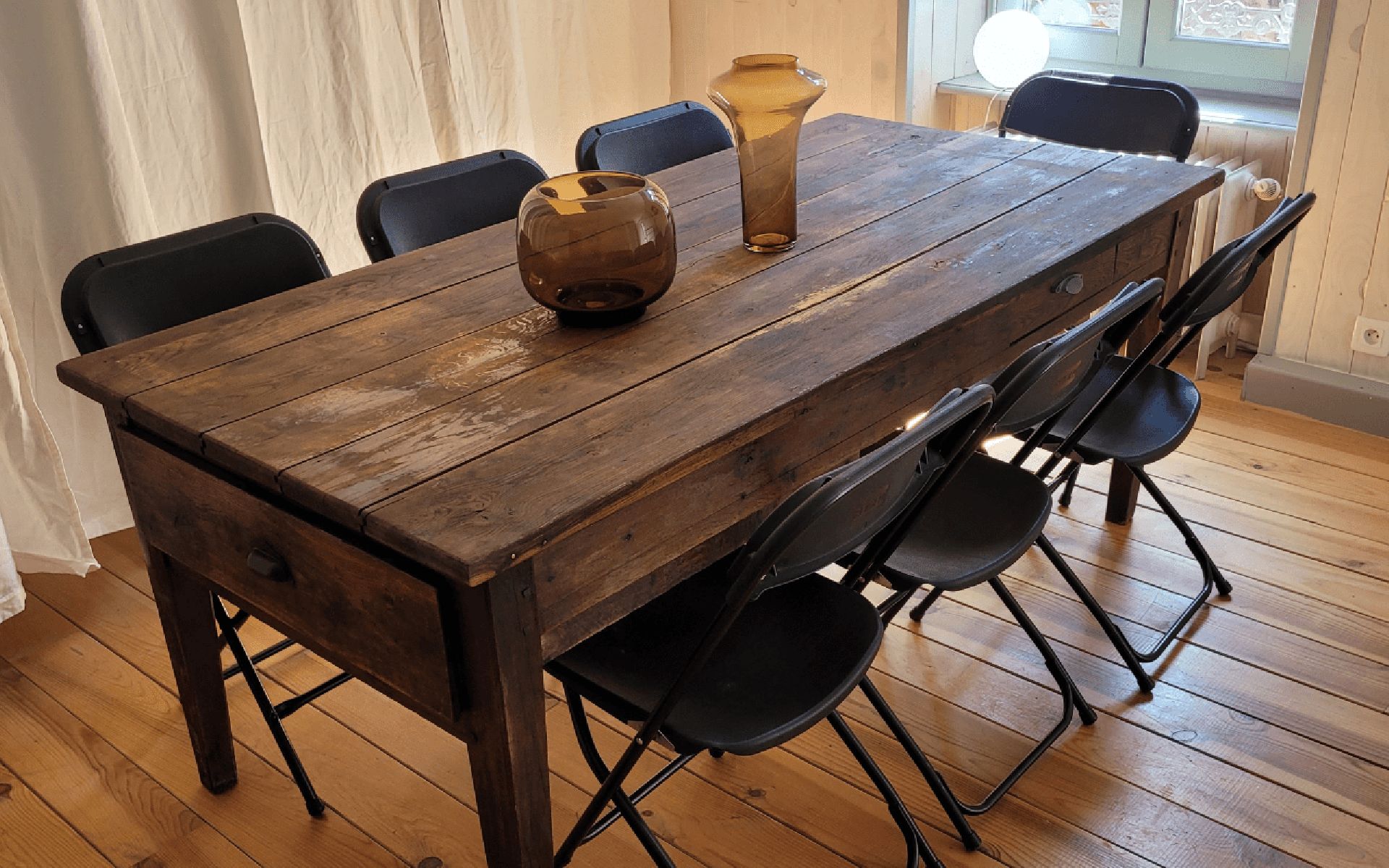 Una mesa de comedor de madera con sillas, un florero y una ventana al fondo.