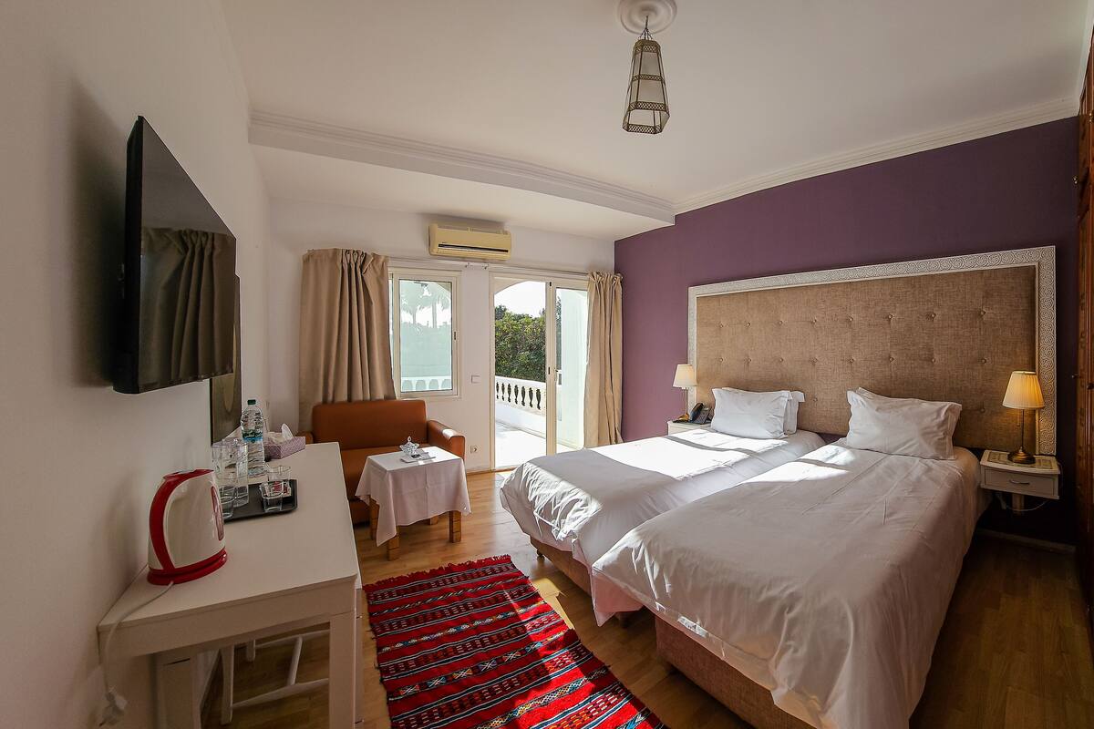 A hotel room with a large bed, a bedside table with a lamp, a small table with a telephone, a chair, and a view of the outdoors through a sliding door.