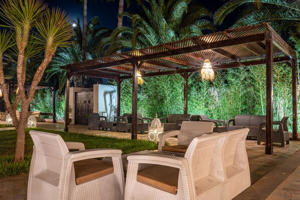 Un espacio al aire libre con mobiliario de mimbre bajo una pérgola de madera, rodeado de abundante follaje y palmeras.