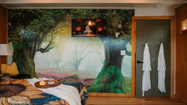 Un dormitorio con un mural grande de un bosque, una cama con cubrecama floral, un televisor montado en la pared y una puerta con toallas colgadas.