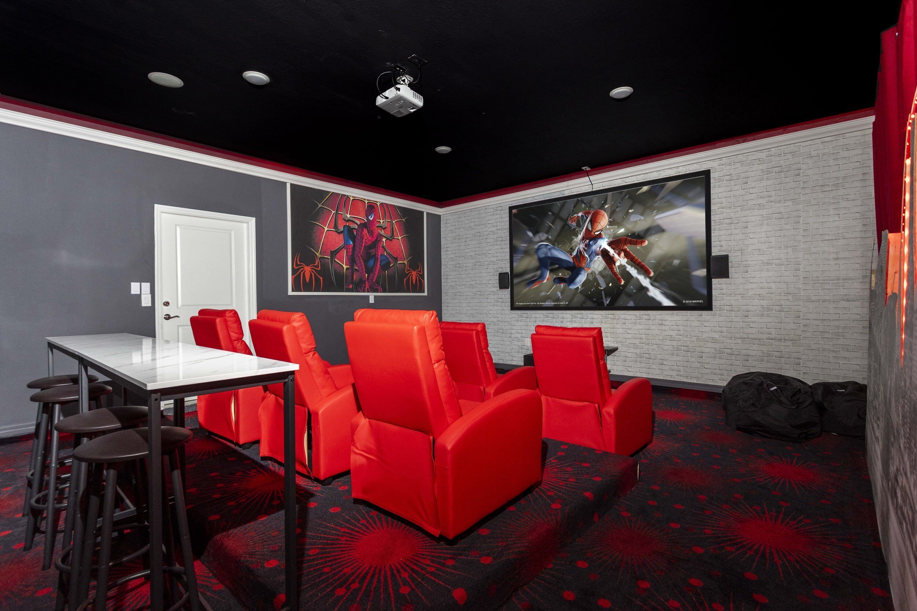 A home theater room with a white brick wall, red and black patterned carpet, a white table, red chairs, and two large posters on the wall.