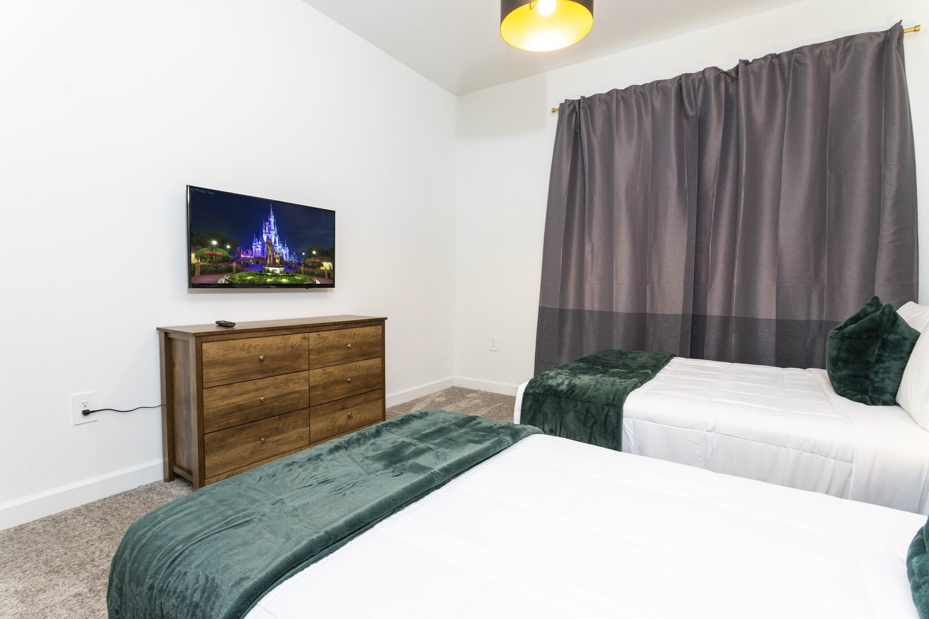 Une chambre avec deux lits, une commode en bois, un téléviseur fixé au mur et une fenêtre avec des rideaux.