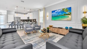 A modern living room with a large flat-screen TV, grey sofas, and a dining area with bar stools.