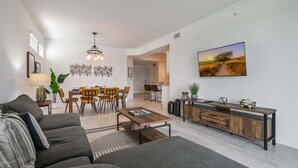 Una sala moderna con sofá, una mesa de centro, comedor con sillas, un televisor montado en la pared y una cocina visible al fondo.