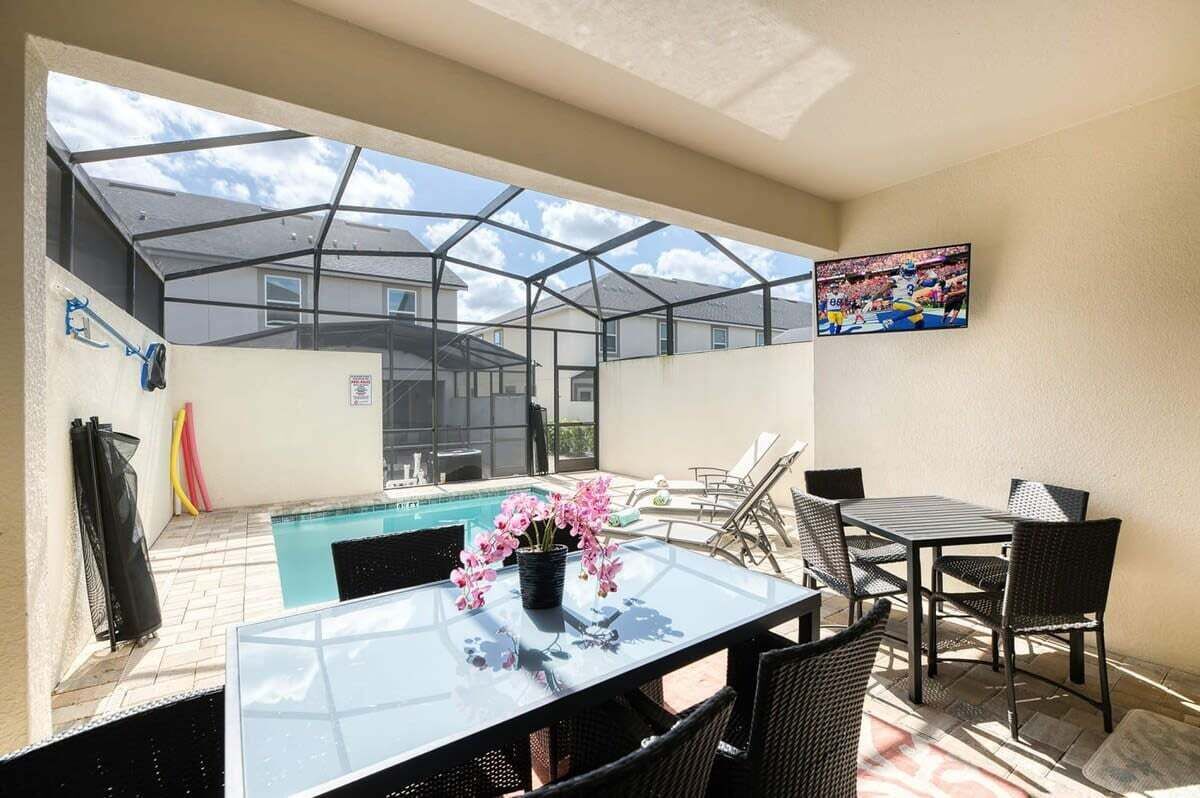 A glass-enclosed patio with a pool, outdoor furniture, and a TV mounted on the wall.