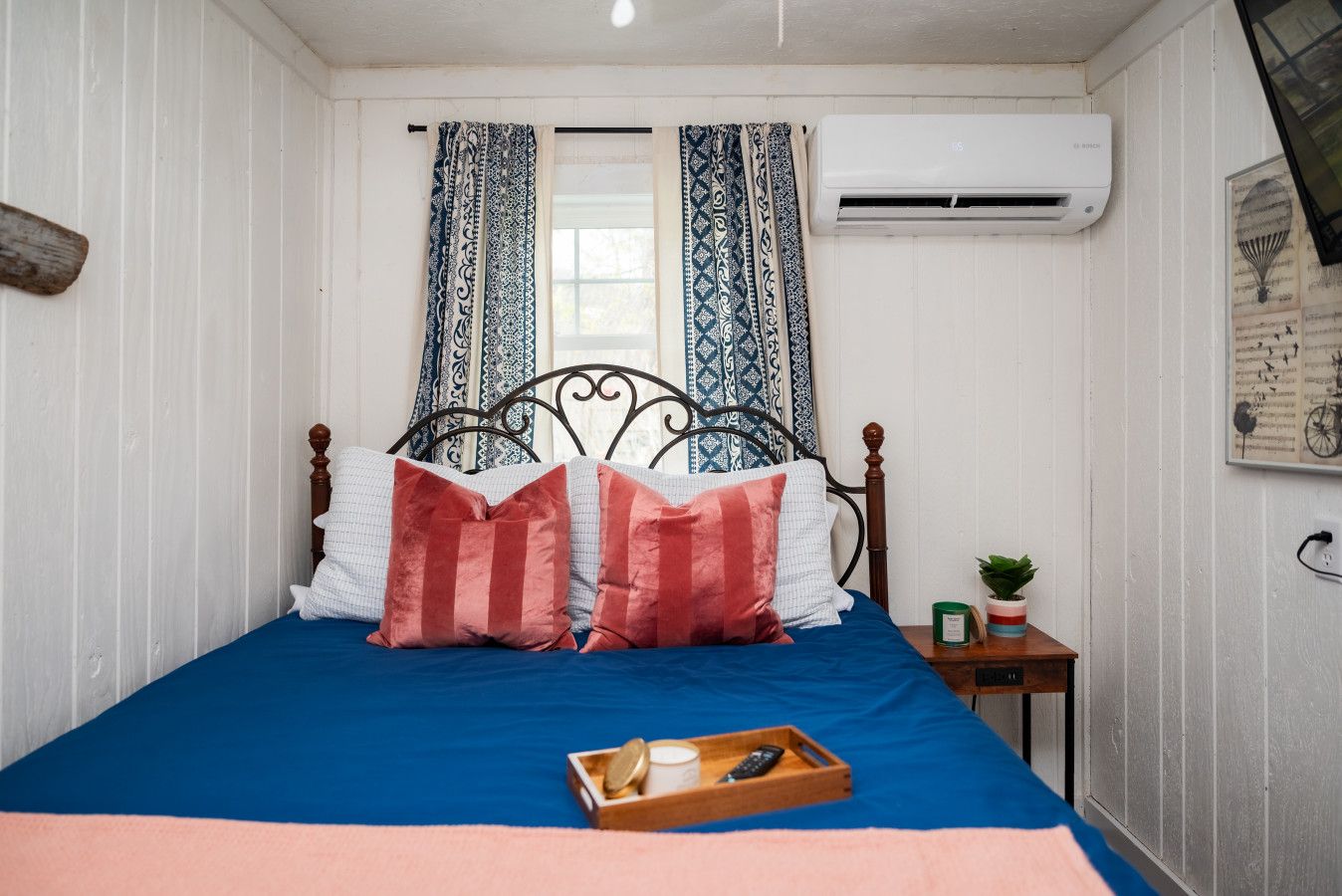 A bedroom with a bed, curtains, an air conditioner, a small table with a plant, and a tray with a cup and remote control.