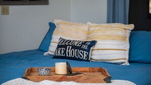 A bed with blue bedding and pillows, a wooden tray with a candle, a mug, and a sign that reads "Welcome to the Lake House".
