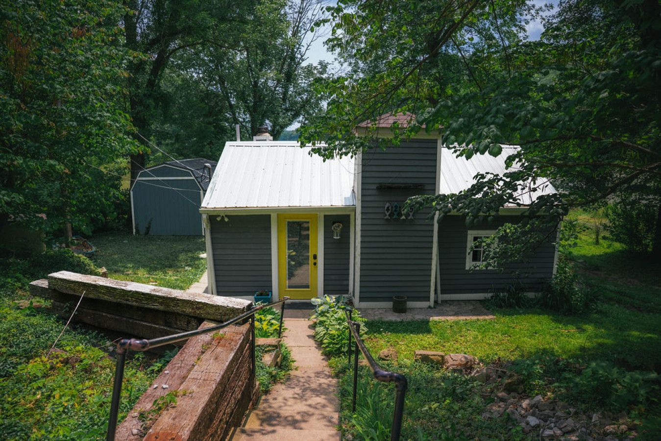 Une petite maison à un seul étage, avec une porte jaune, entourée d’une végétation luxuriante et un chemin de pierres menant à l’entrée.
