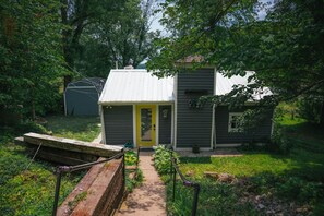 A small, single-story house with a yellow door, surrounded by lush greenery and a stone pathway leading to the entrance.