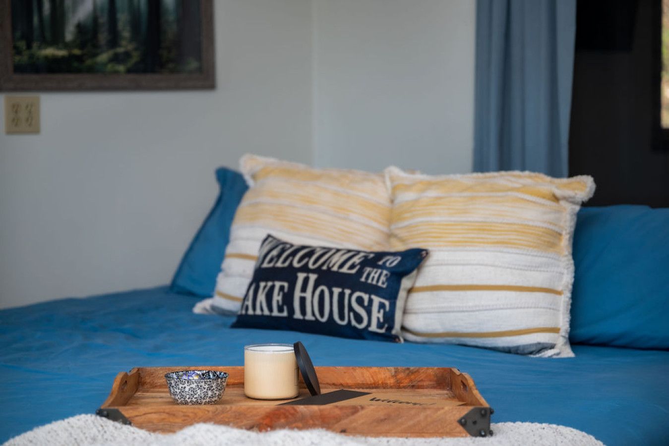 A bed with blue bedding and pillows, a wooden tray with a candle, a mug, and a sign that reads "Welcome to the Lake House".