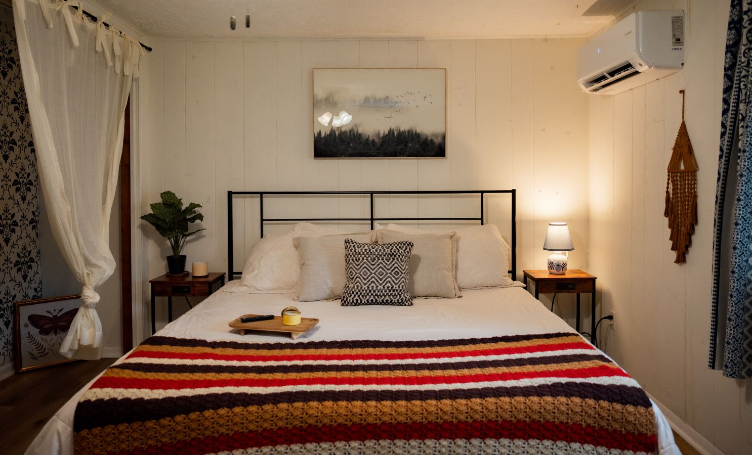 Un lit bien fait, recouvert d’une couverture à rayures, une table de chevet avec une lampe, un tableau encadré au mur et une fenêtre avec des rideaux.