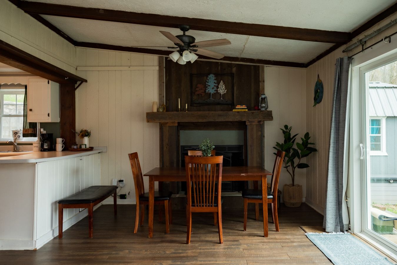 Uma área de jantar com mesa e cadeiras de madeira, um ventilador de teto e vista para o exterior através de uma janela.