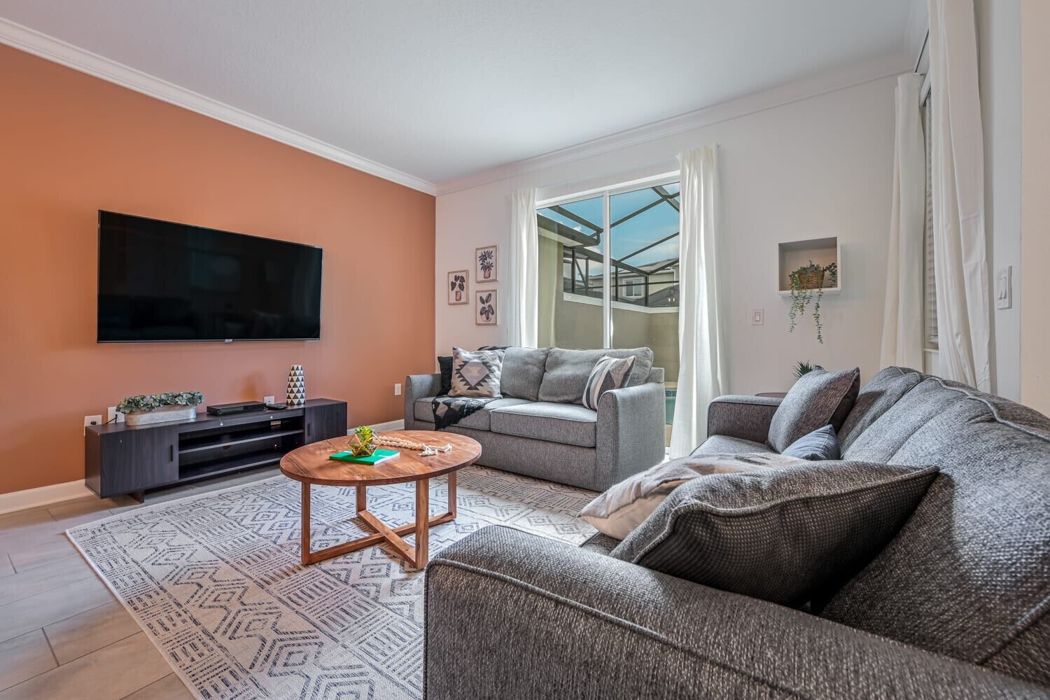 Una sala de estar moderna con una televisión de pantalla plana, sofás grises, una mesa de centro y un gran ventanal con cortinas.