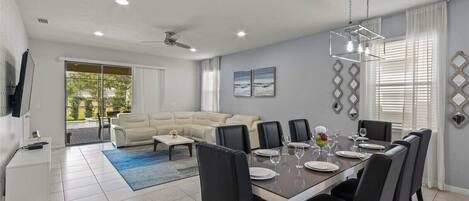 A modern dining area with a large table set for six, surrounded by black chairs. There is a sofa and a television in the background, and the room has a ceiling fan and recessed lighting.