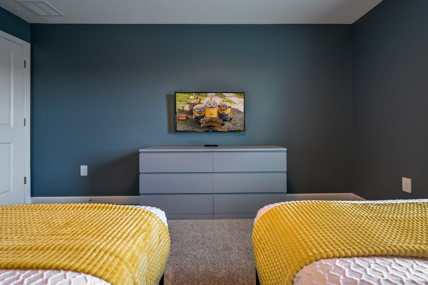 A bedroom with two beds, a dresser, and a wall-mounted TV displaying a Minions movie.