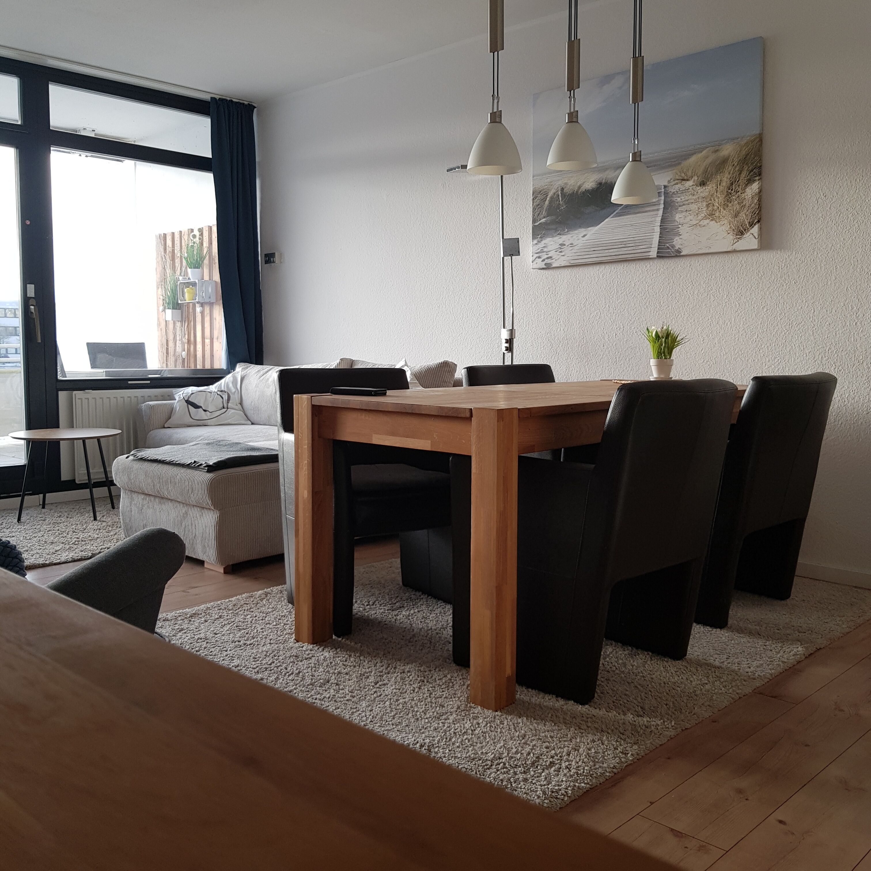 A modern living room with a wooden dining table, black chairs, a sofa, and a large window with curtains.