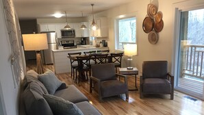 Una sala de estar moderna con un sofá gris, dos sillones, piso de madera y una cocina al fondo.