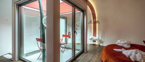 Habitación de hotel moderna con un amplio ventanal corredero que da a un balcón, una cama con cabecera roja y una pequeña zona de estar con una silla roja y una mesa blanca.