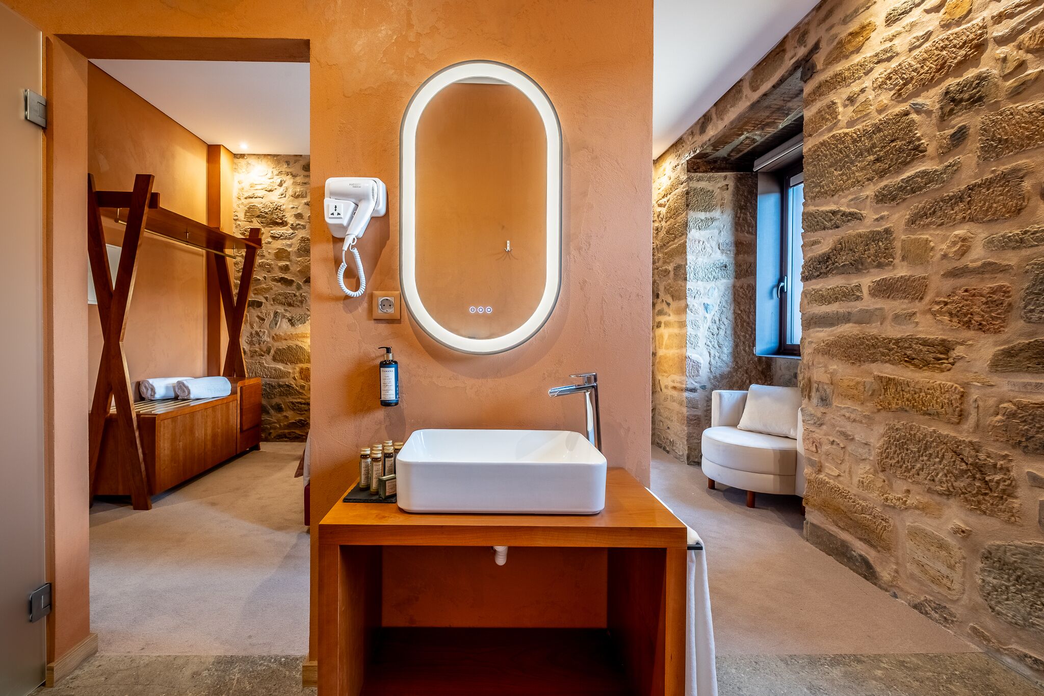 A bathroom with a wooden vanity, a large oval mirror, a hairdryer, and a stone wall.