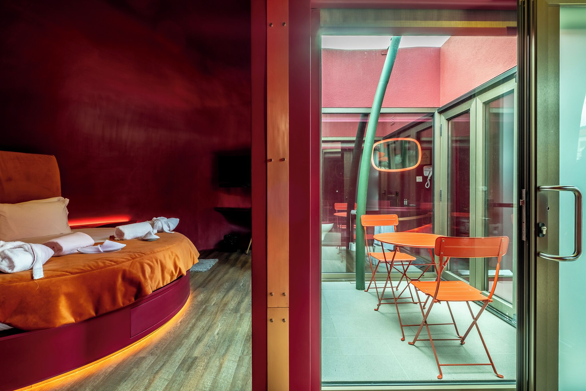 A modern hotel room with a circular bed, a glass door leading to a balcony with orange chairs and a table.