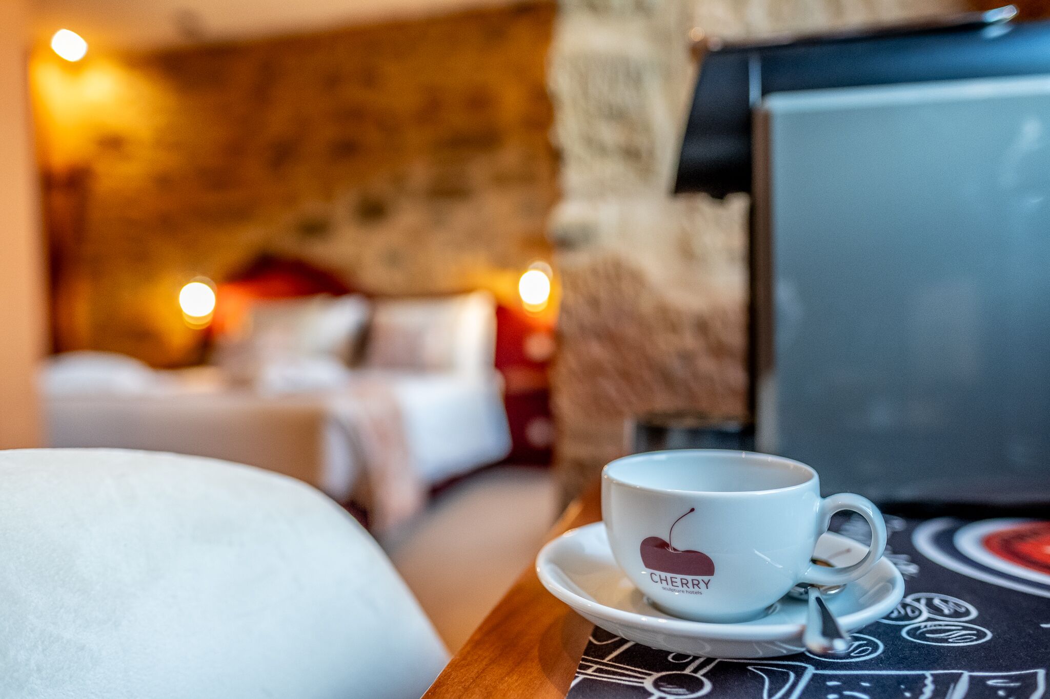 A cup of coffee on a table with a saucer, a bed, and a stone wall in the background.