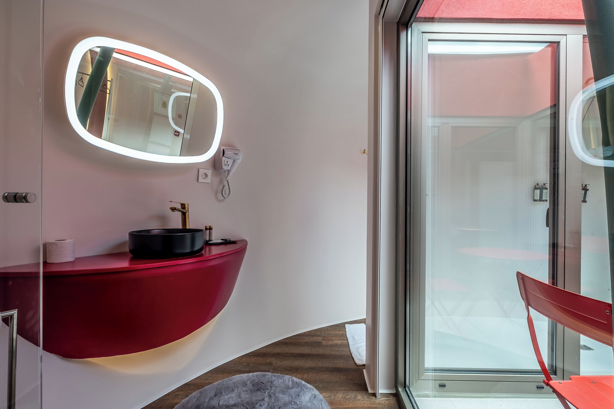 A modern bathroom with a curved red vanity, a round mirror with integrated lighting, a glass shower enclosure, and a red chair.