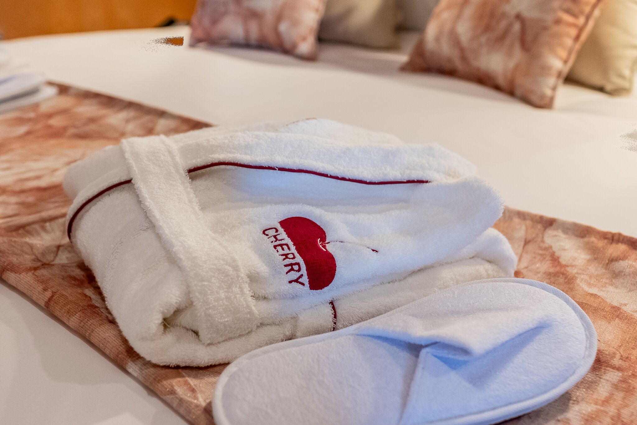 A neatly folded white bathrobe with a red logo on a wooden tray, accompanied by a pair of white slippers.