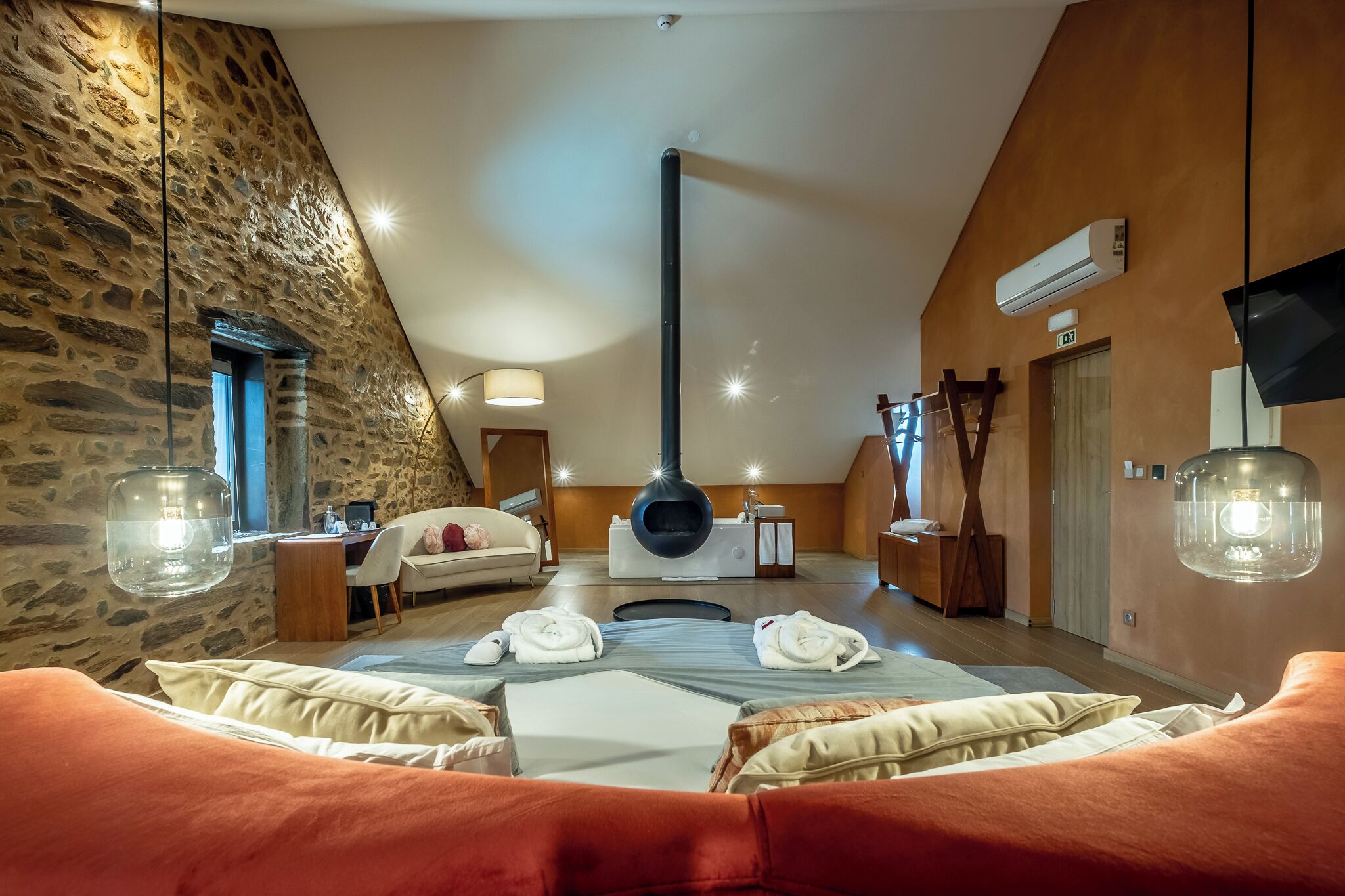 Ein geräumiger Raum mit Steinwand, Kamin, einem Bett mit orangefarbener Bettwäsche und einem Schreibtisch mit Lampe.