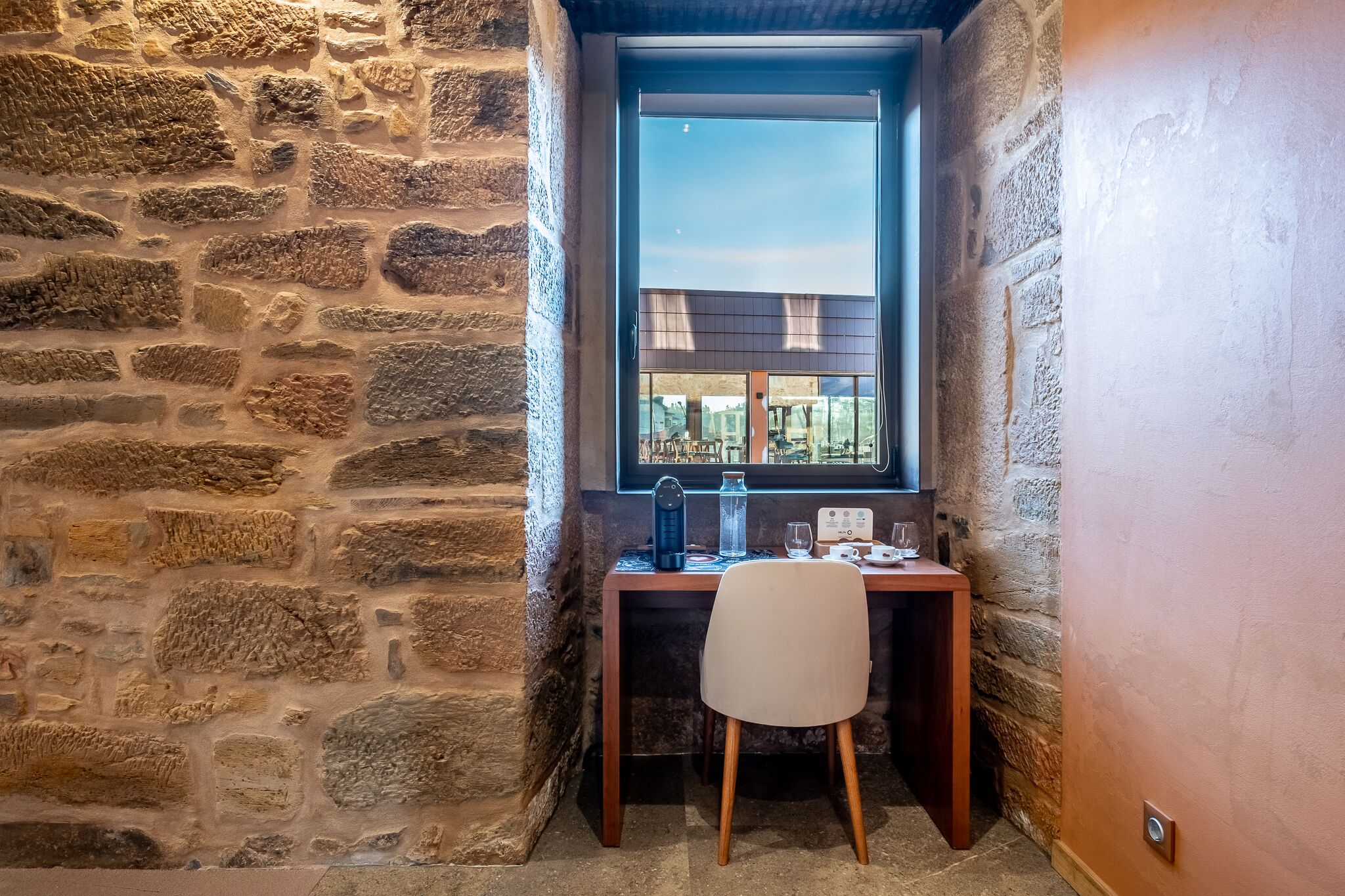 A corner room with a stone wall, a wooden desk, a chair, and a window with a view of buildings.