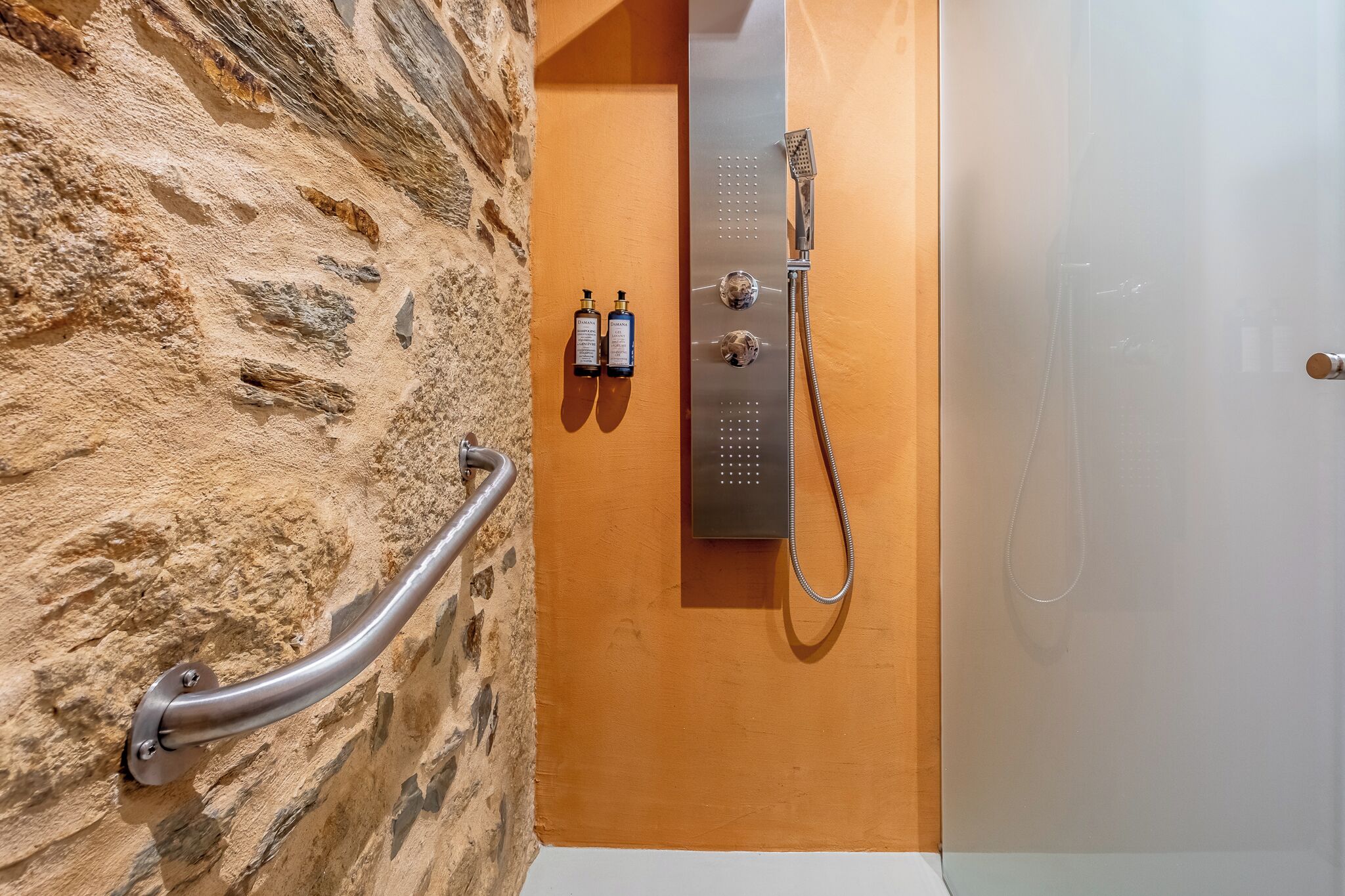 A bathroom with a stone wall, a shower with a handheld showerhead, and two bottles of shampoo.