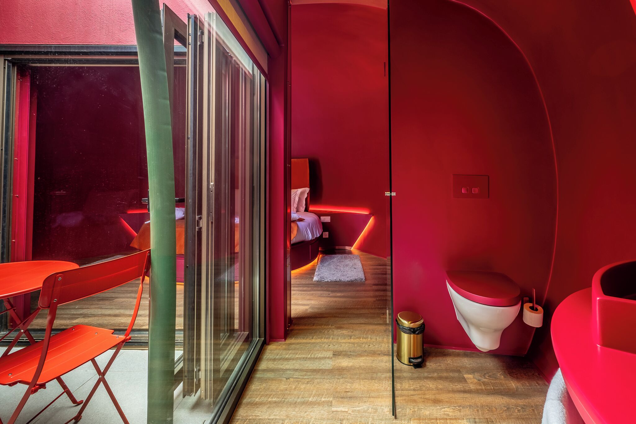 Ein modernes Zimmer mit Glastür zu einem Außenbereich, einem Bett mit roter Akzentwand, einem kleinen Teppich und einer Toilette mit rotem Sitz.