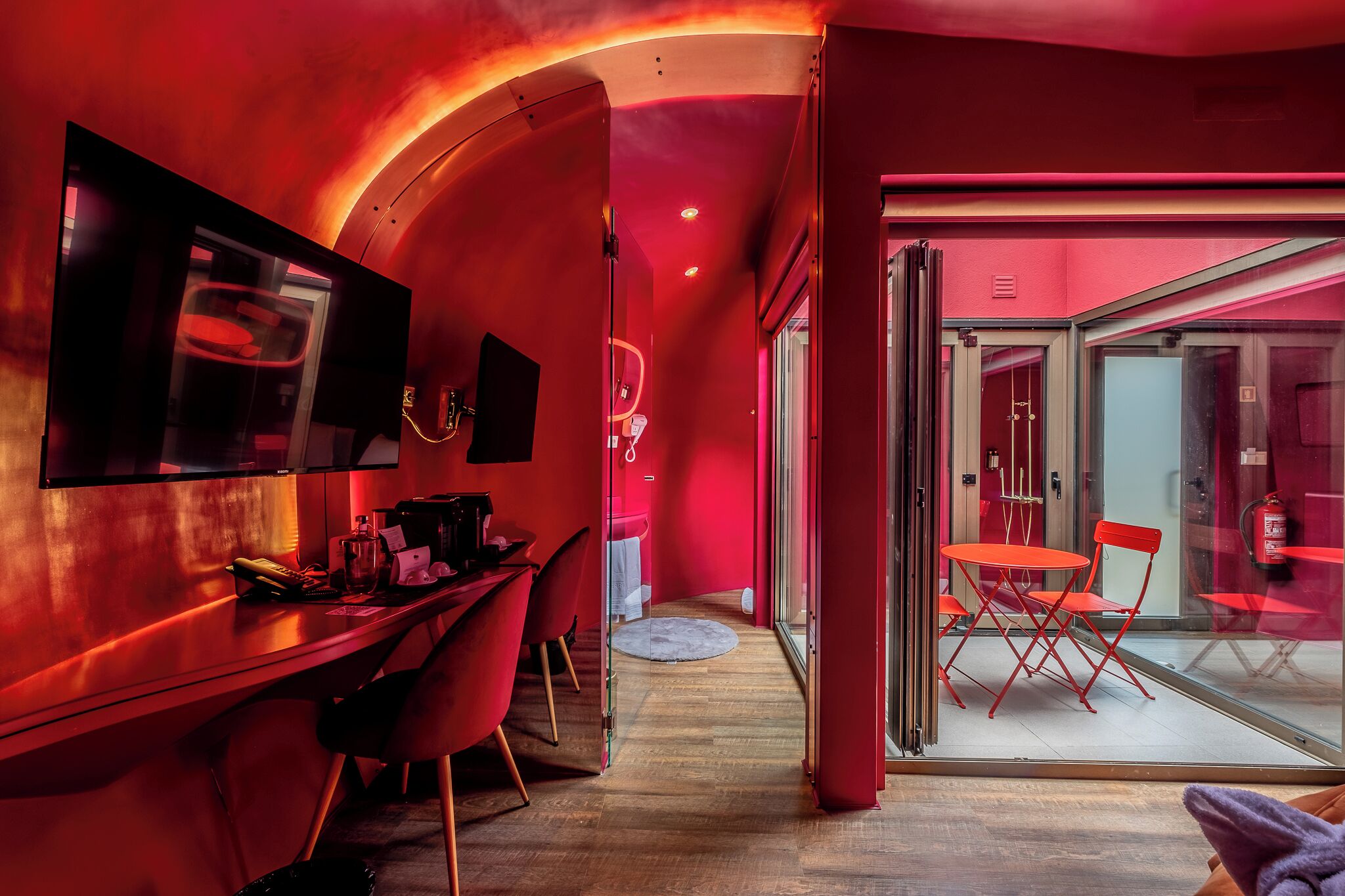 Una habitación de hotel moderna con una pared curva, un escritorio con una silla, un televisor y una puerta de cristal que da a un comedor con sillas rojas y una mesa.