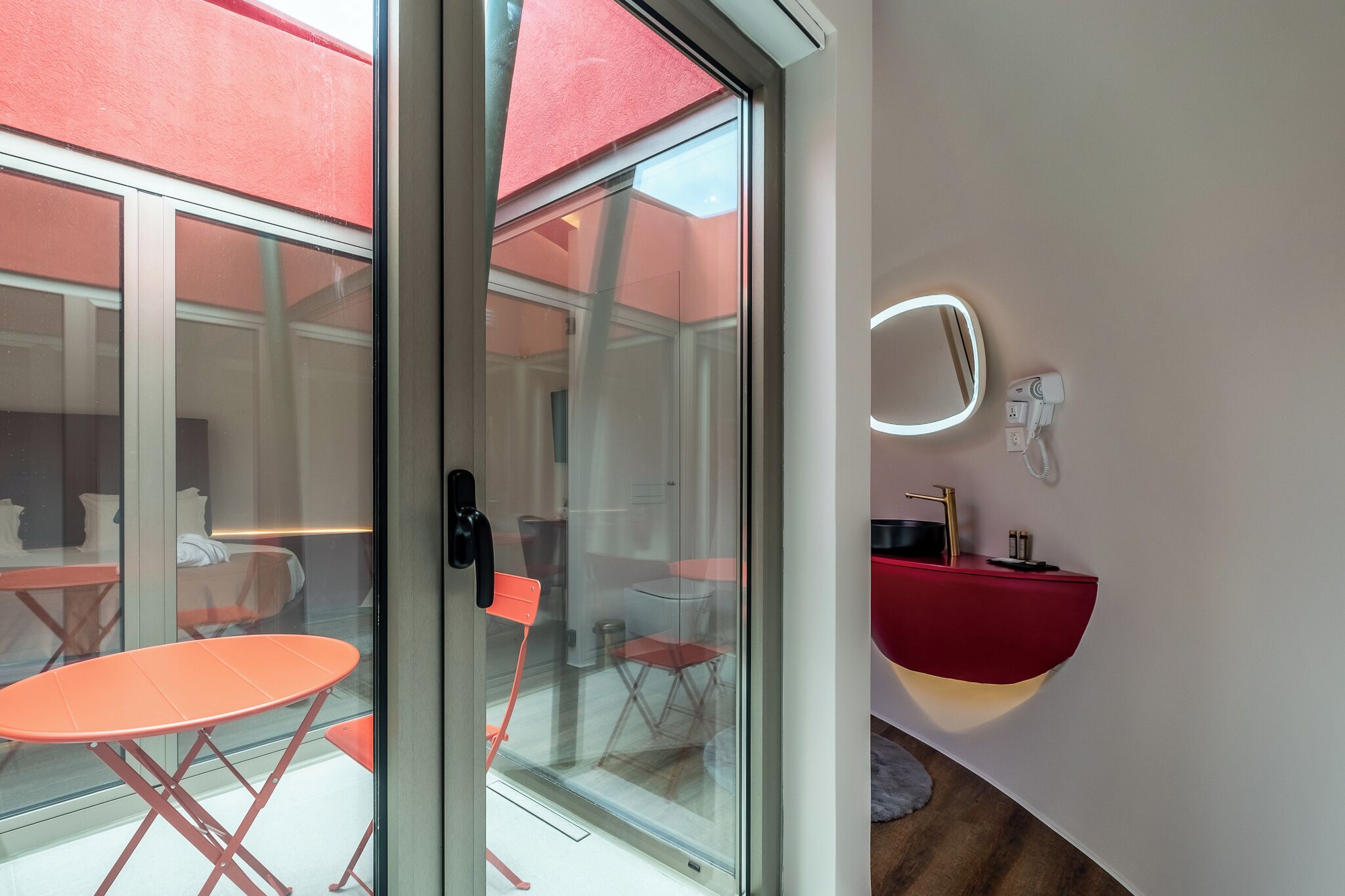 A modern hotel room with a glass door leading to an outdoor seating area, a red sink, and a wall-mounted mirror.