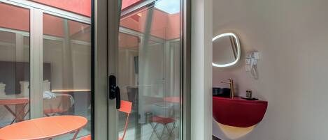 A modern hotel room with a glass door leading to an outdoor seating area, a red sink, and a wall-mounted mirror.