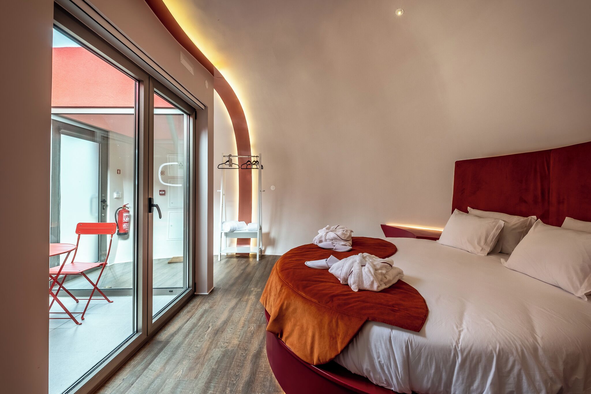 A modern hotel room with a large bed, a red headboard, a glass door leading to a balcony, and a small table with a chair.