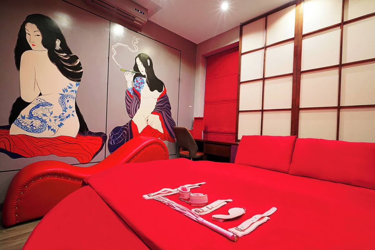 A room with a red bedspread, a red chair, and a wall with two large paintings.