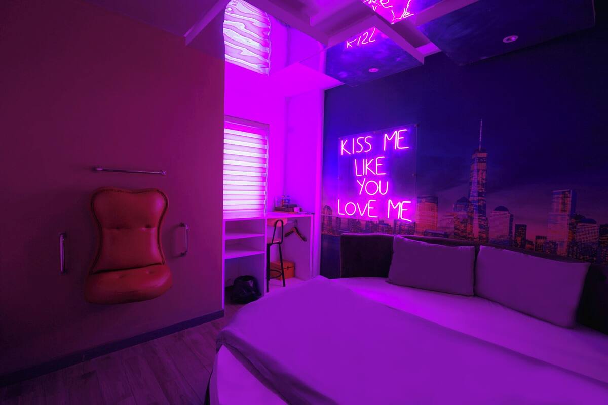 A modern bedroom with a neon sign reading "KISS ME LIKE YOU LOVE ME" and a cityscape view.