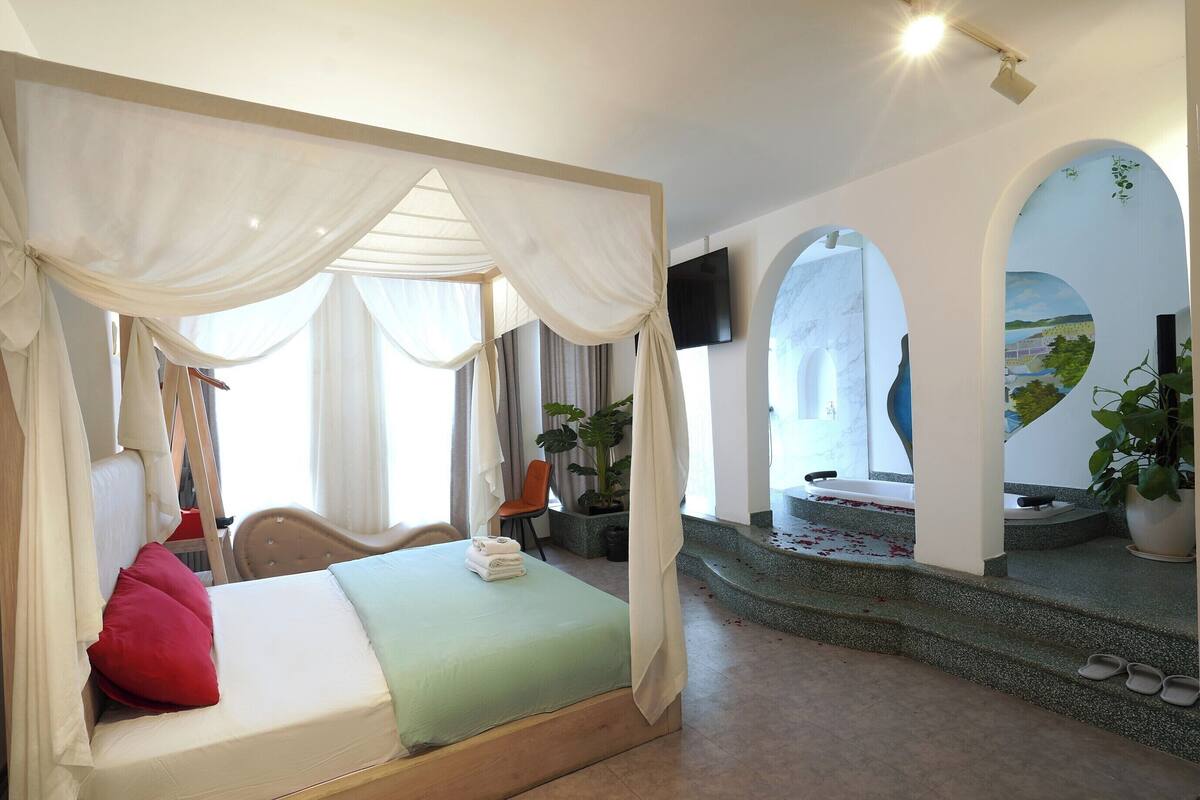 A bedroom with a four-poster bed, a green bedspread, and a red pillow. There is a chair and a plant in the room.