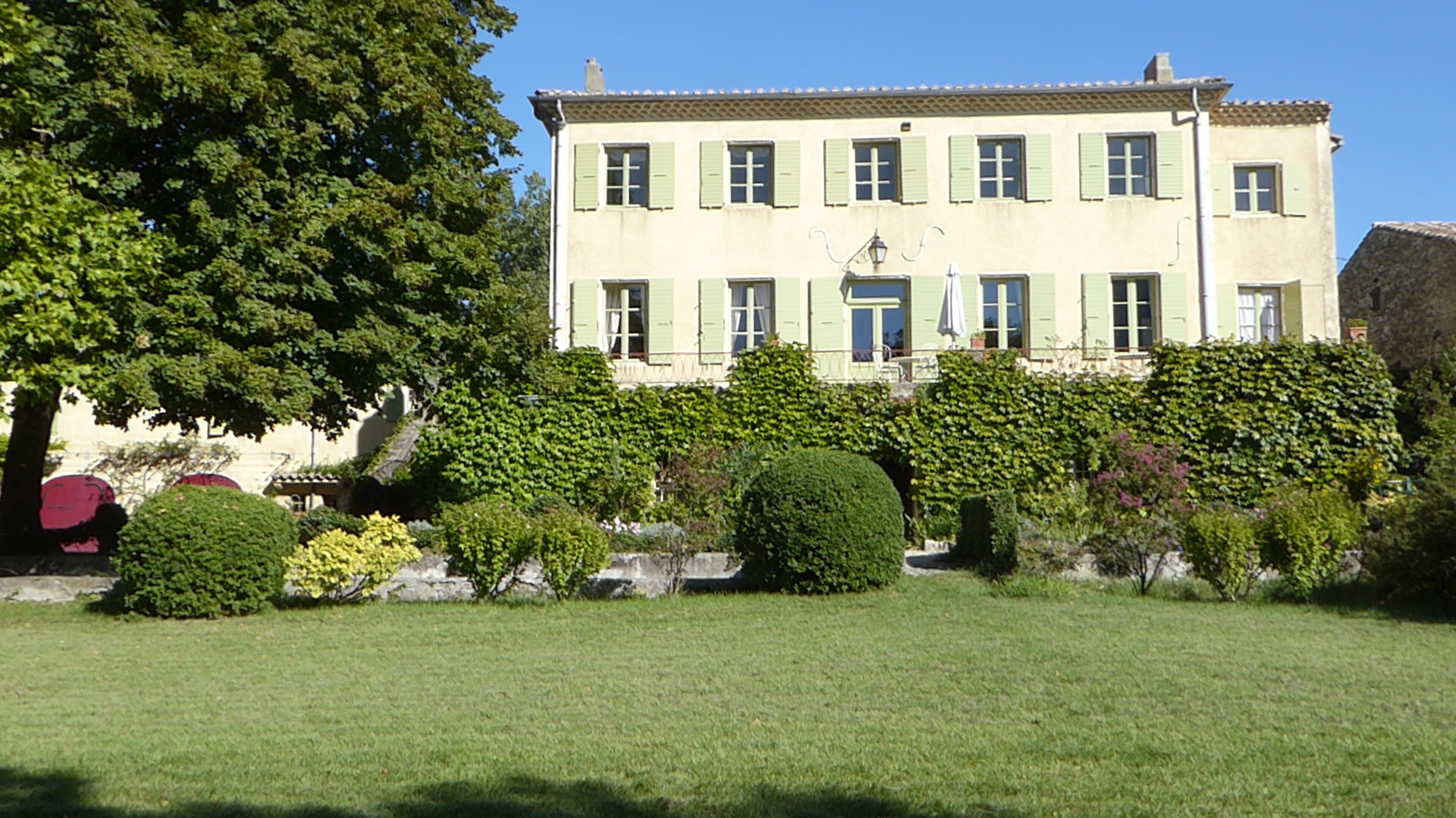 Ein zweistöckiges Gebäude mit einer symmetrischen Fassade, zahlreichen Fenstern mit Fensterläden und einem gepflegten Garten mit Hecken und Rasenfläche.