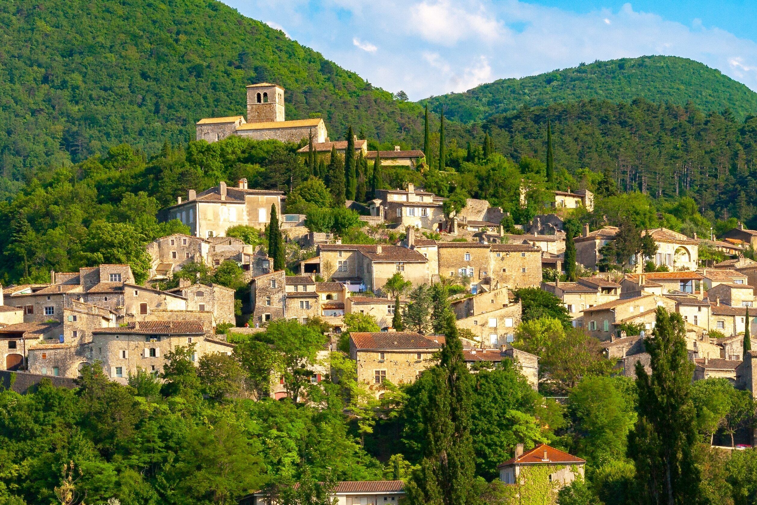 A hillside village with stone buildings, a church with a bell tower, and lush greenery.
